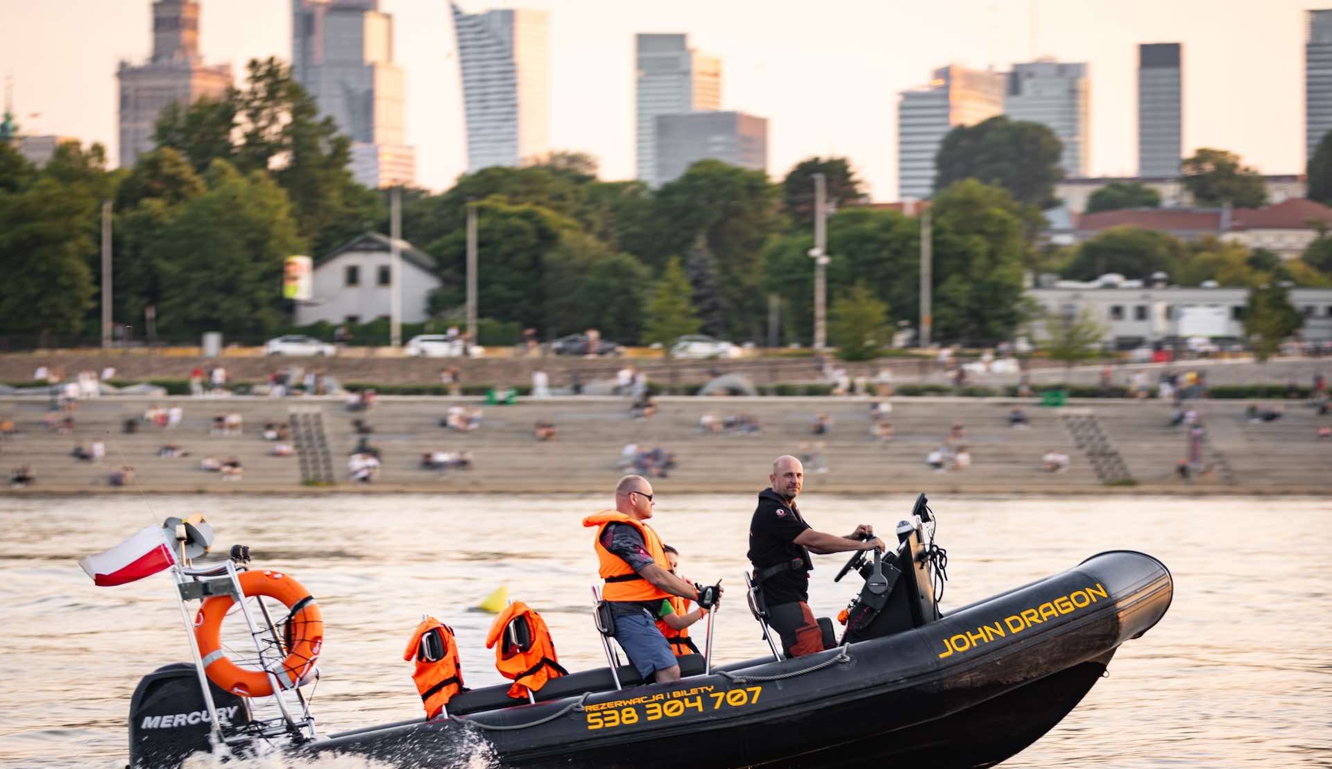 Motorówka RIB John Dragon wykonująca zwrot na Wiśle, w tle drapacze chmur centrum Warszawy i plaża z ludźmi